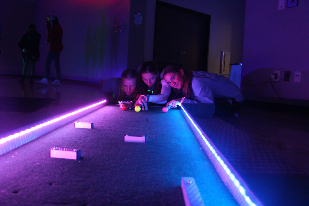 three teenage girls in the colored glow of a darkened room playing pool shuffleboard