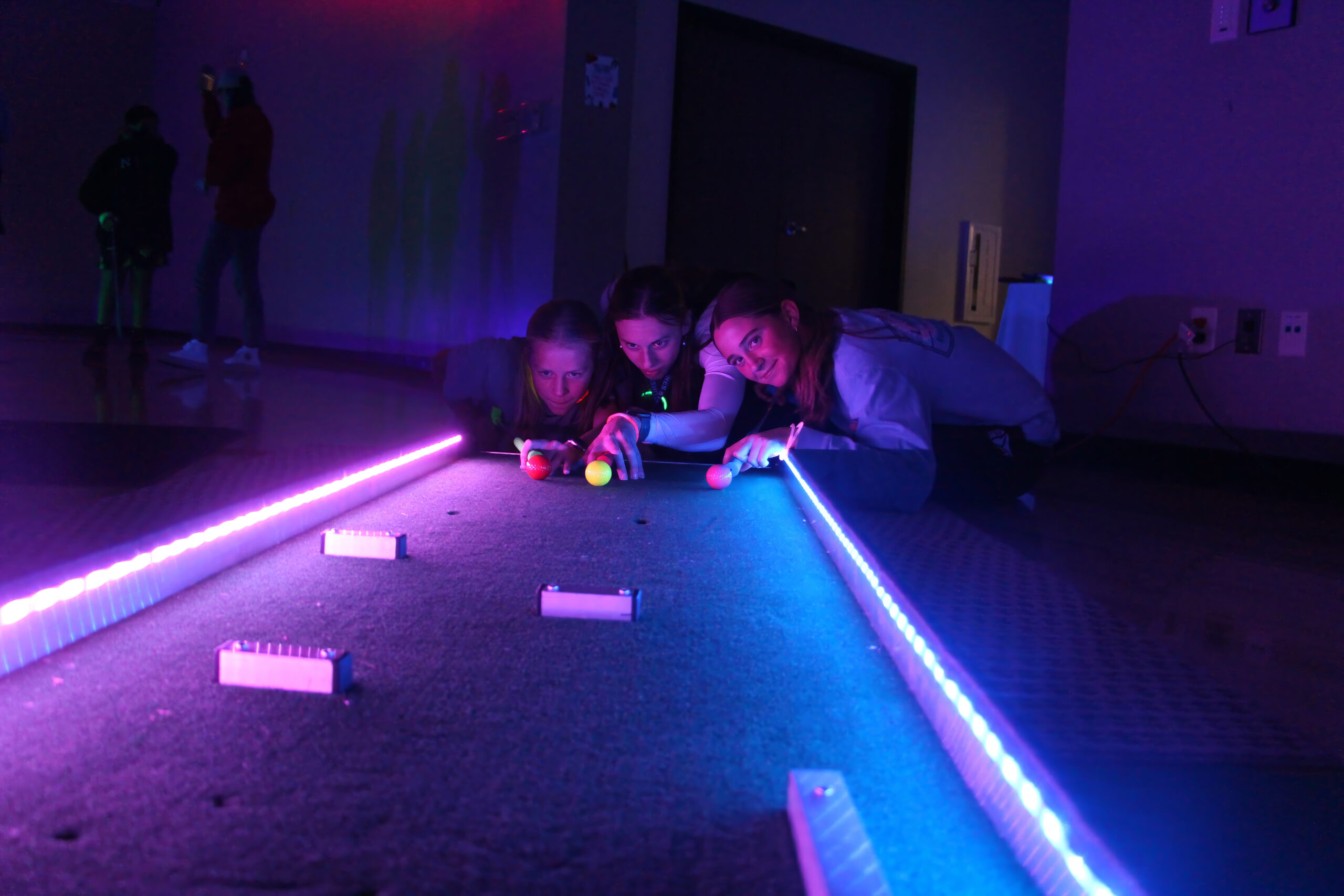 three teenage girls in the colored glow of a darkened room playing pool shuffleboard